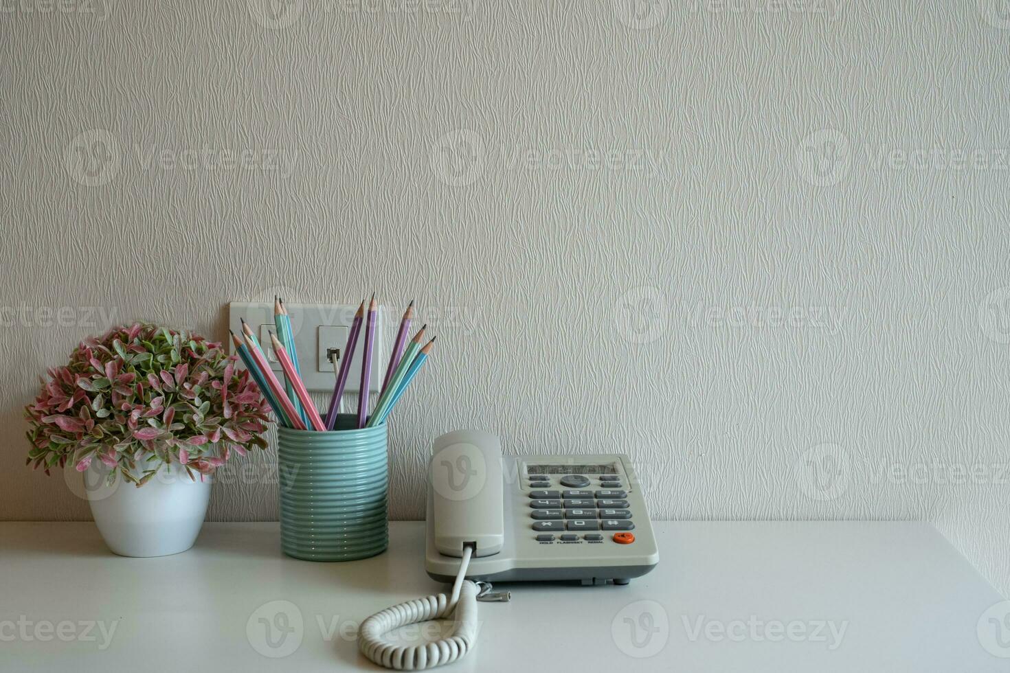 Telephone and pastel pencils in the blue glass on the desk at gray wall background photo