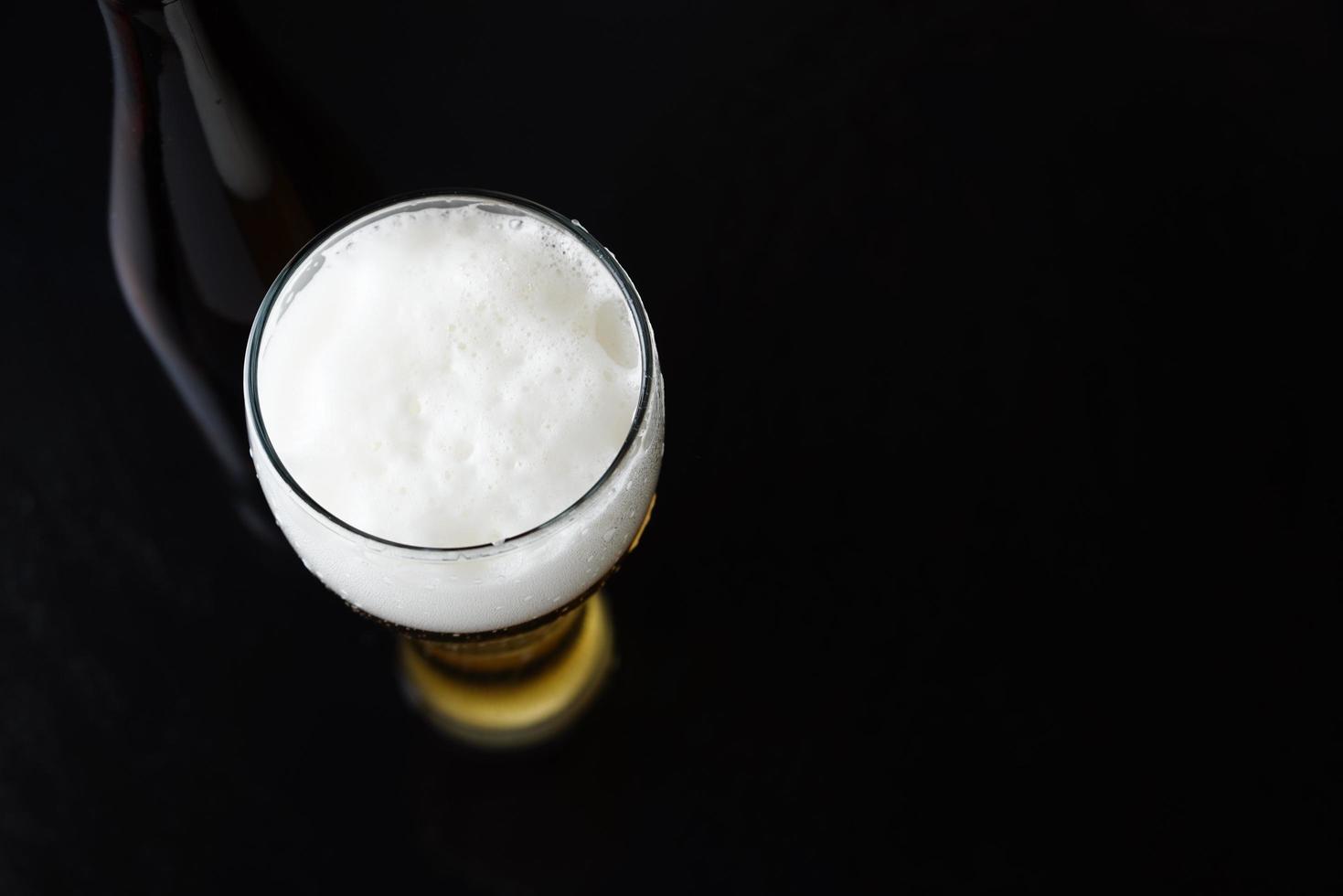 un vaso de cerveza fría con espuma y una botella al fondo. fondo oscuro negro con espacio de copia. foto