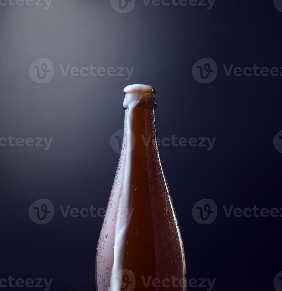 Front view of a bottle of cold beer with overflowing foam against blue grunge vintage background. Organic photography, vertical, square image style. photo