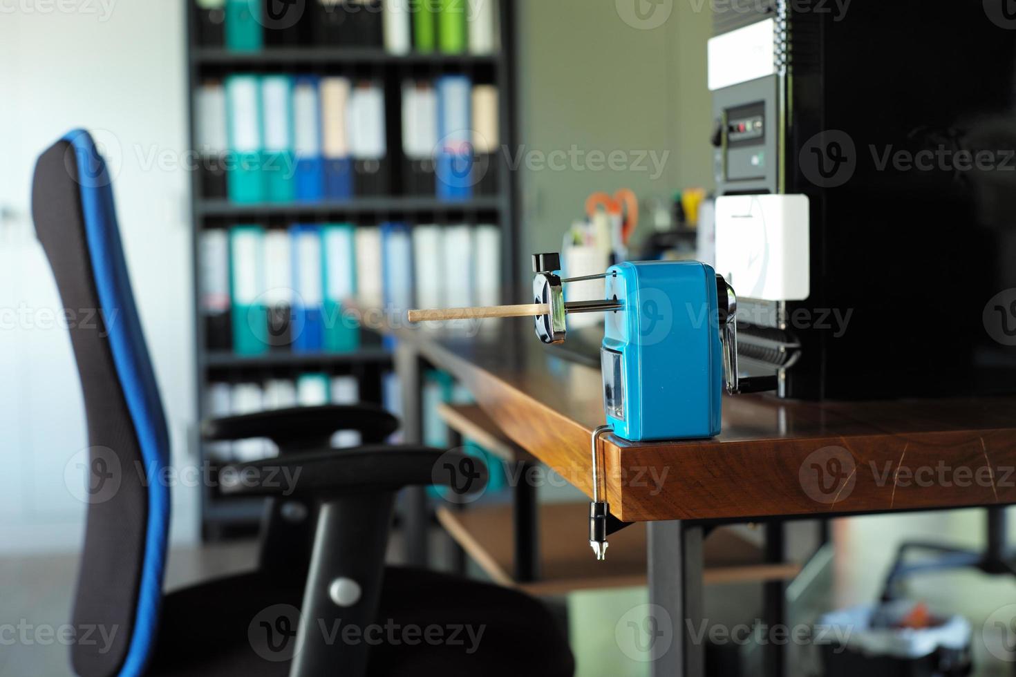 Closeup pencil sharpener with pencil inside installed on the top of wooden table with blurred computer and stationary in background photo