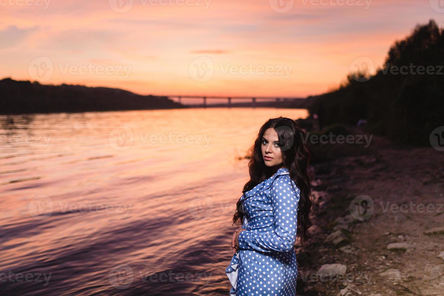 Beautiful young girl with long dark wavy hair standing at the bank of the river photo