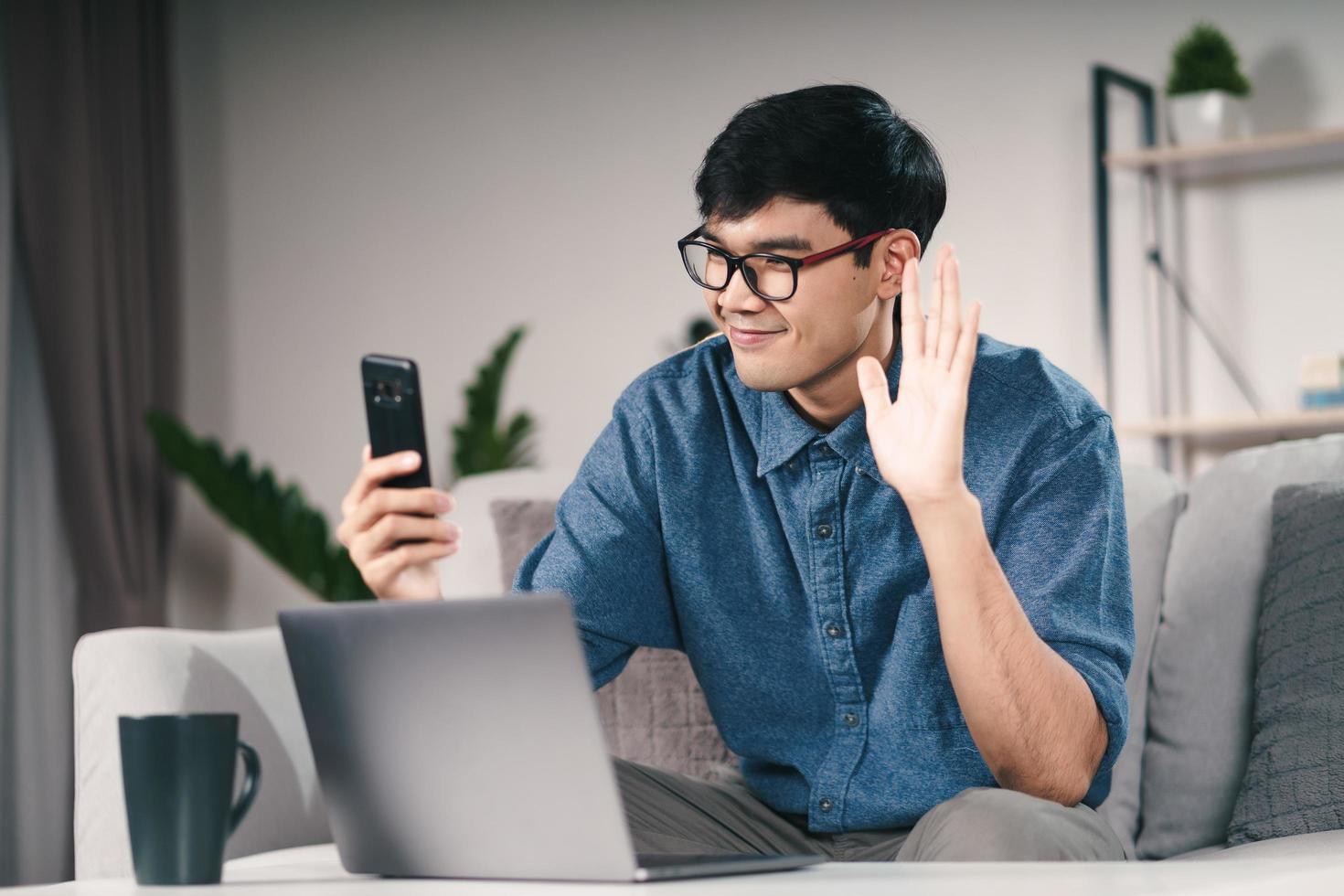 Joven asiático con teléfono inteligente para llamada de videoconferencia en línea agitando la mano haciendo gesto de saludo en el sofá de la sala de estar por la noche. foto