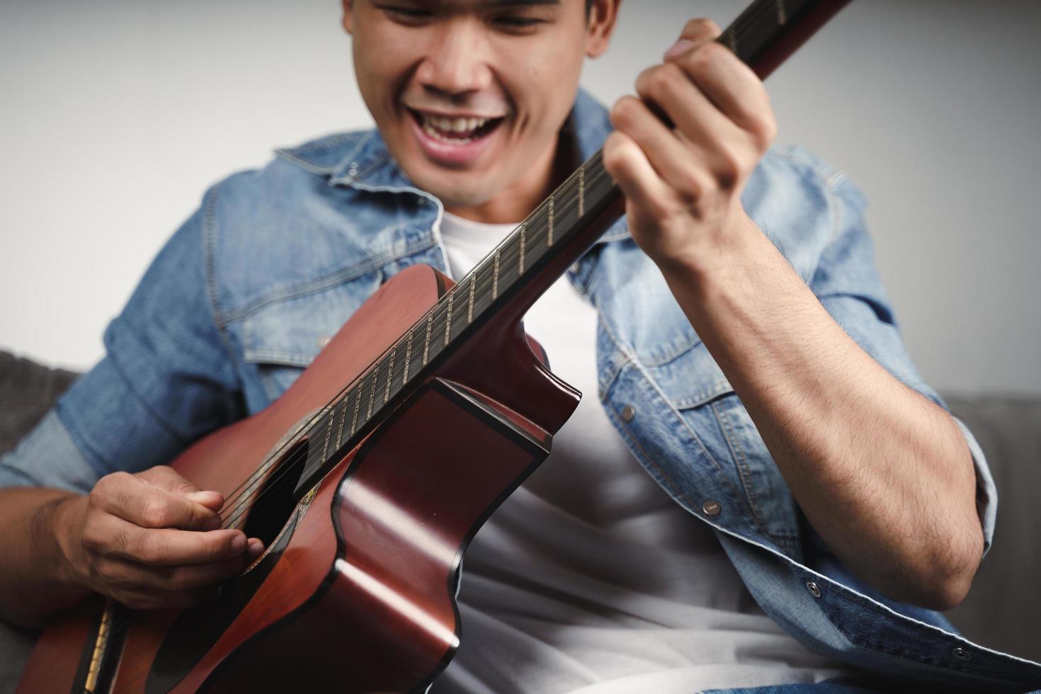 Disfrute de un apuesto hombre asiático practicando o tocando la guitarra en el sofá de la sala de estar foto