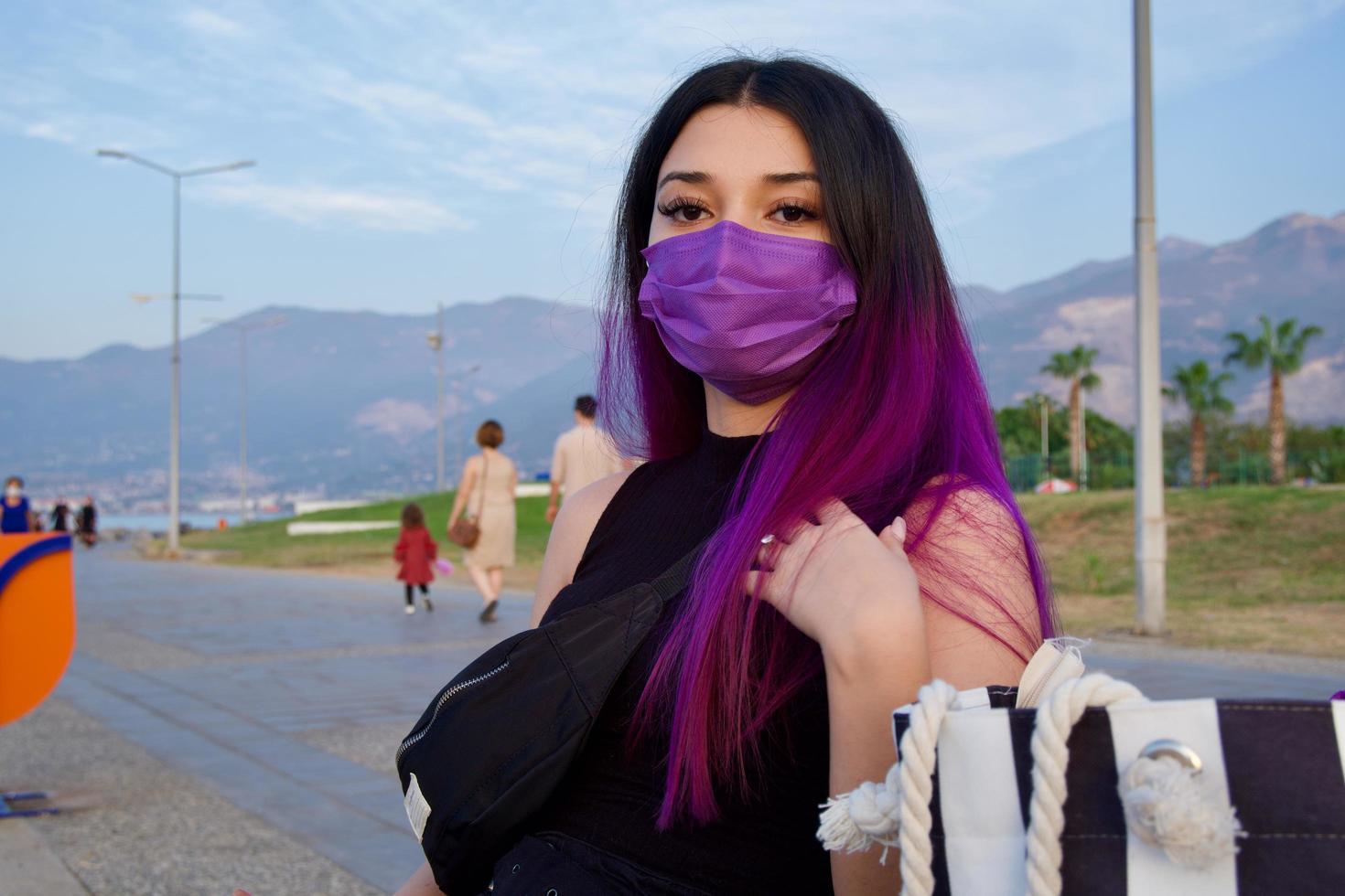 una mujer de cabello púrpura con bolsas de la compra. mujer joven de compras está descansando sentado en el parque. ella usa una máscara púrpura en su rostro debido al covid 19. foto