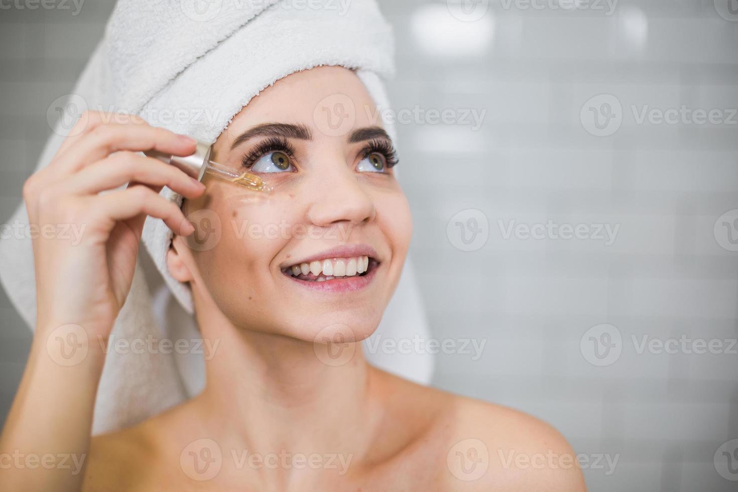 Young woman in white towel apply moisturising oil on face skin. photo