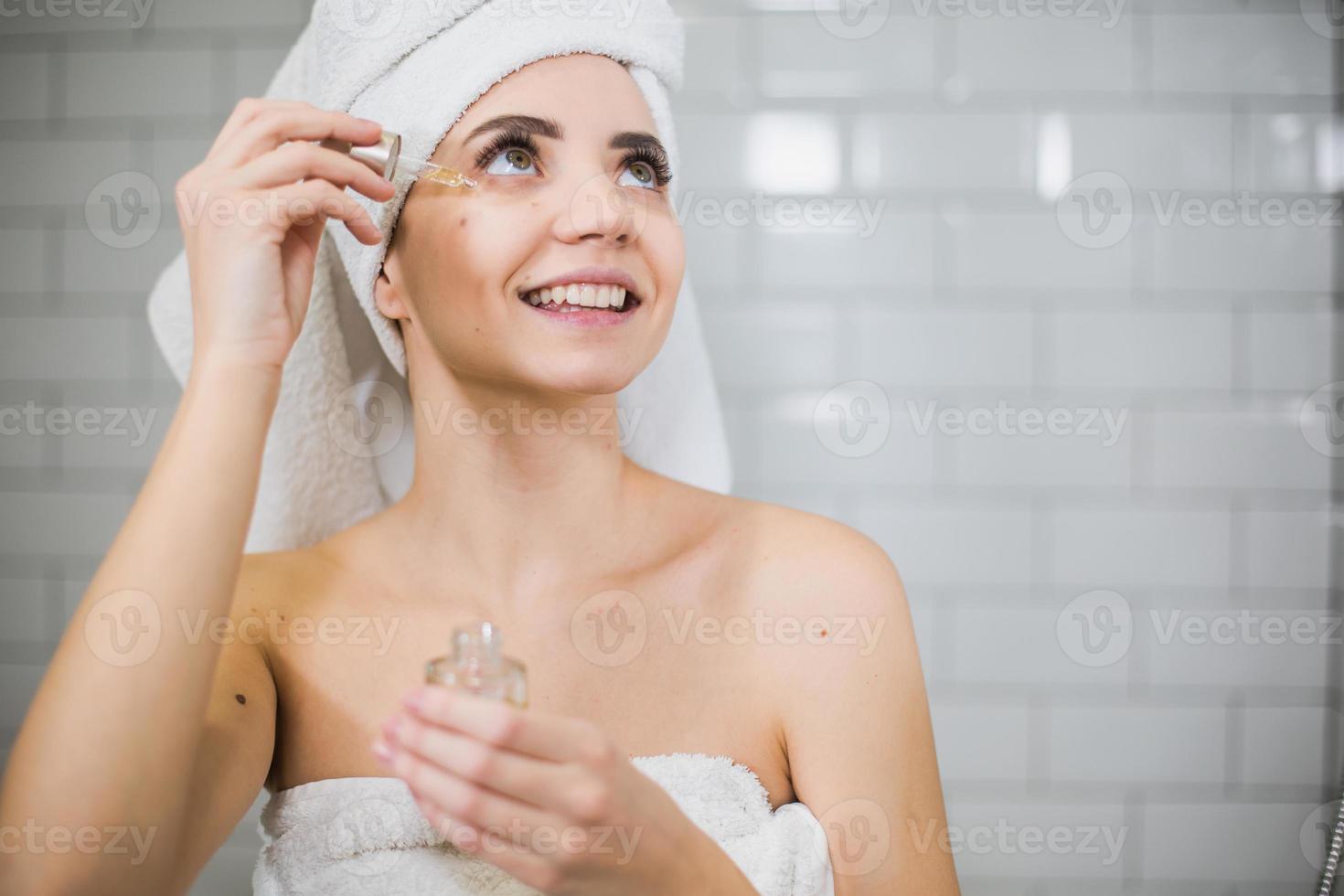 mujer joven en toalla blanca aplicar aceite hidratante en la piel de la cara. foto