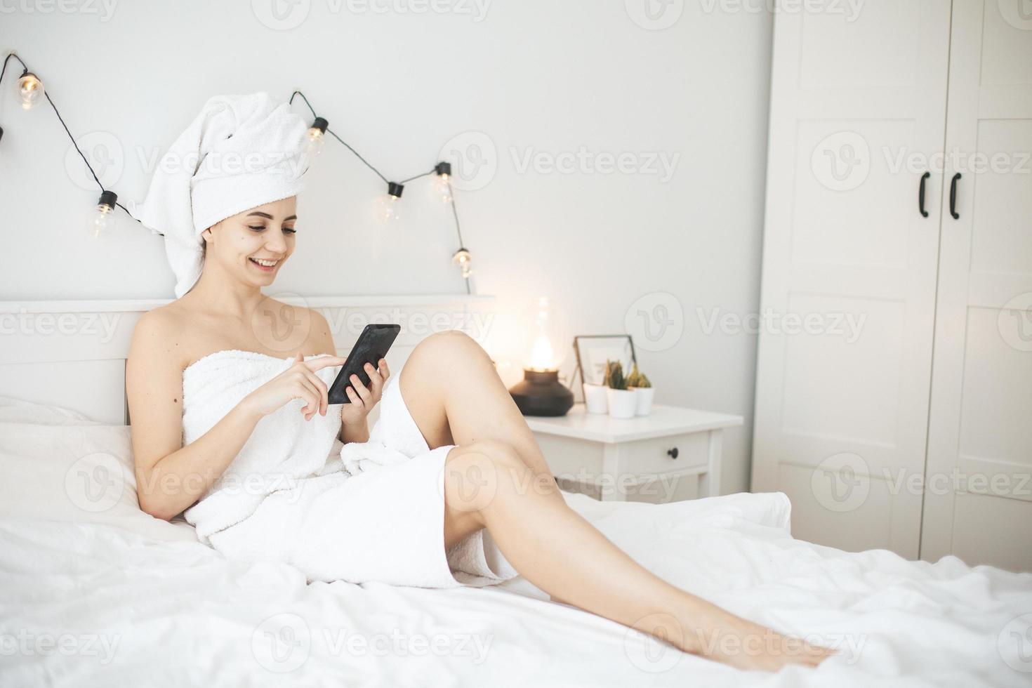 Young woman with towel in white bed texting message on smartphone. photo