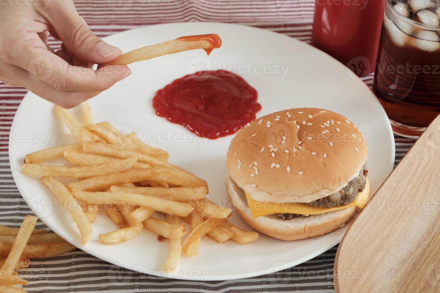 Close-up shot, selective focus. Man's hand was eating French fried, dipping ketchup on a white plate on red tablecloth with hamburgers and cola. Eating junk food or fast food for lunch is unhealthy. photo