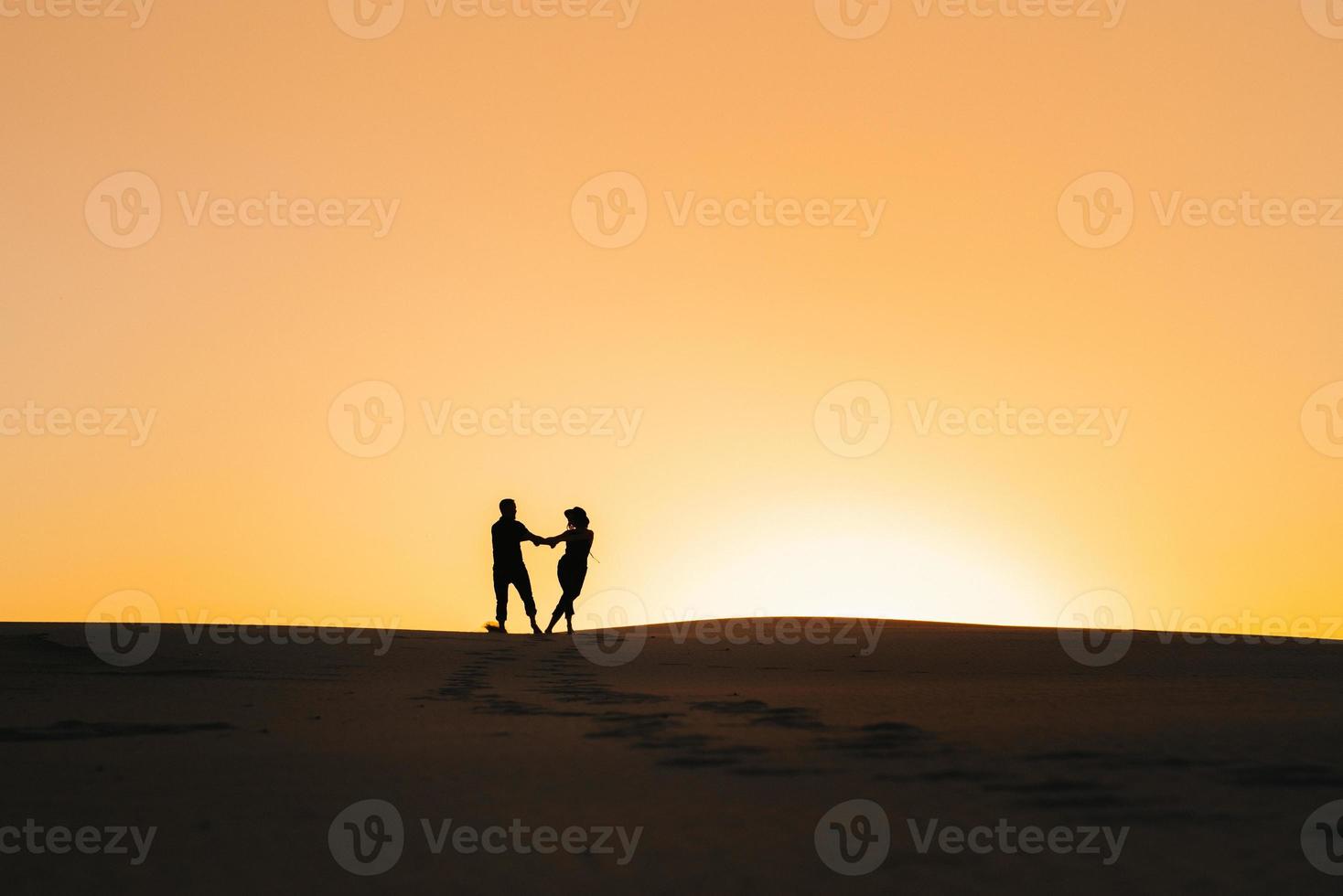 Siluetas de una joven pareja feliz sobre un fondo de naranja atardecer en el desierto de arena foto