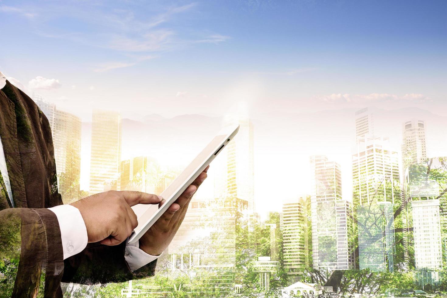 Double exposure of success businessman using tablet with city landscape background. Nature and Building construction. Mixed media photo
