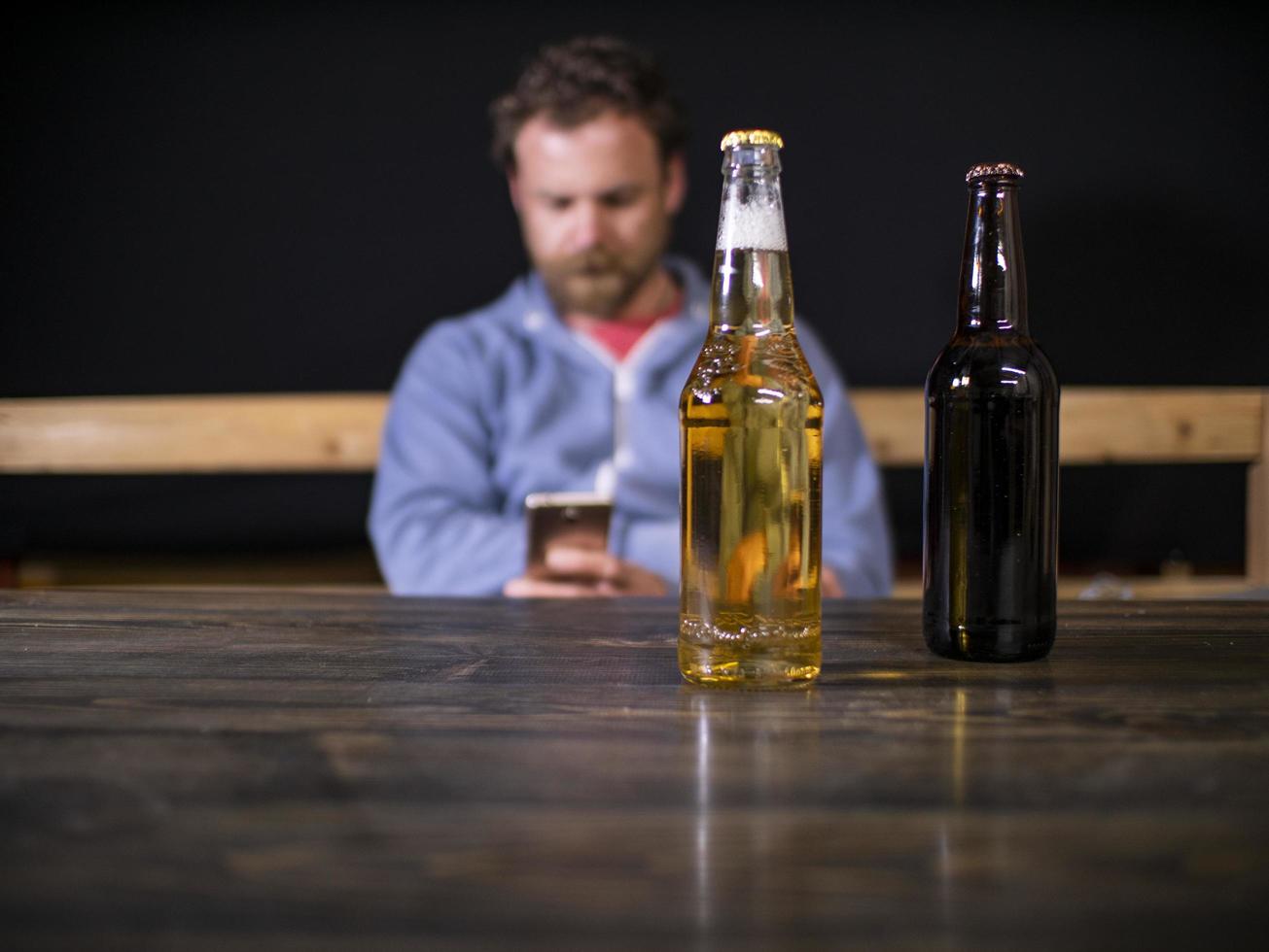 Dos botellas de cerveza están de pie sobre la mesa con el telón de fondo de un hombre sentado que mira por teléfono foto