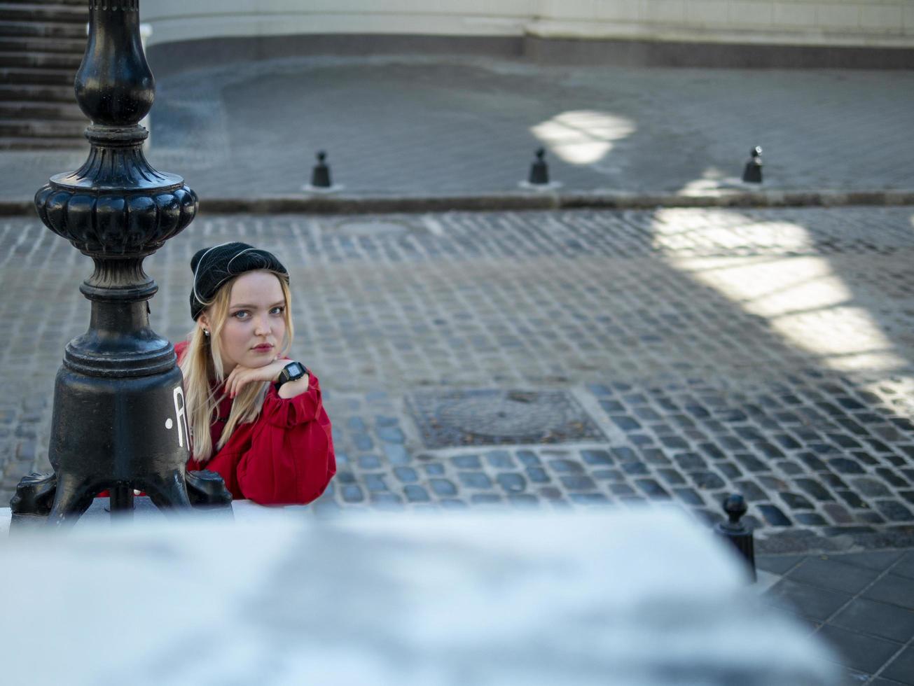 a young girl blonde in a knitted black hat and a red jacket stands near the lantern on the background of a paving stone photo