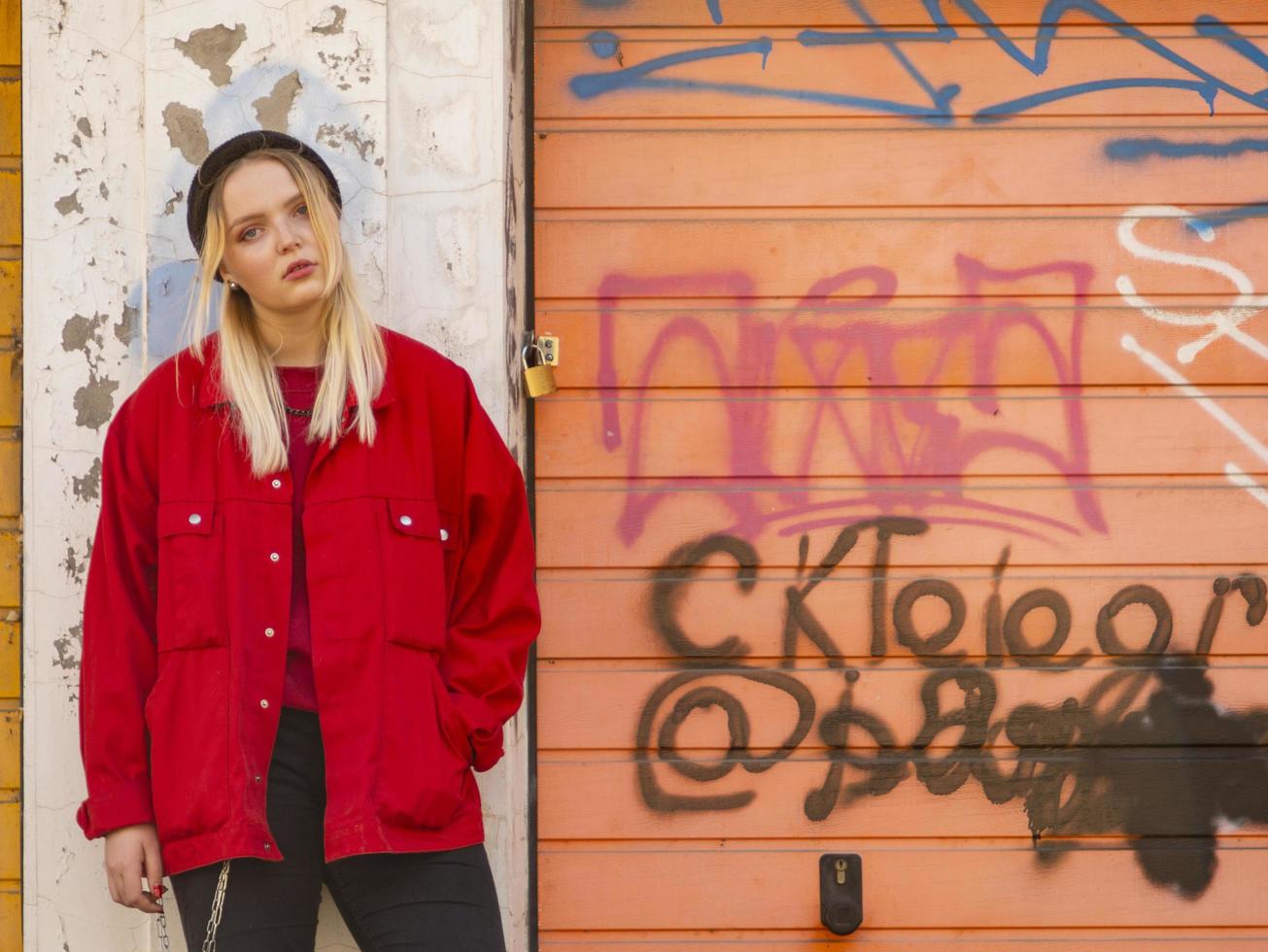 a young girl in a knitted hat and a red jacket stands against the painted wall of a garage photo