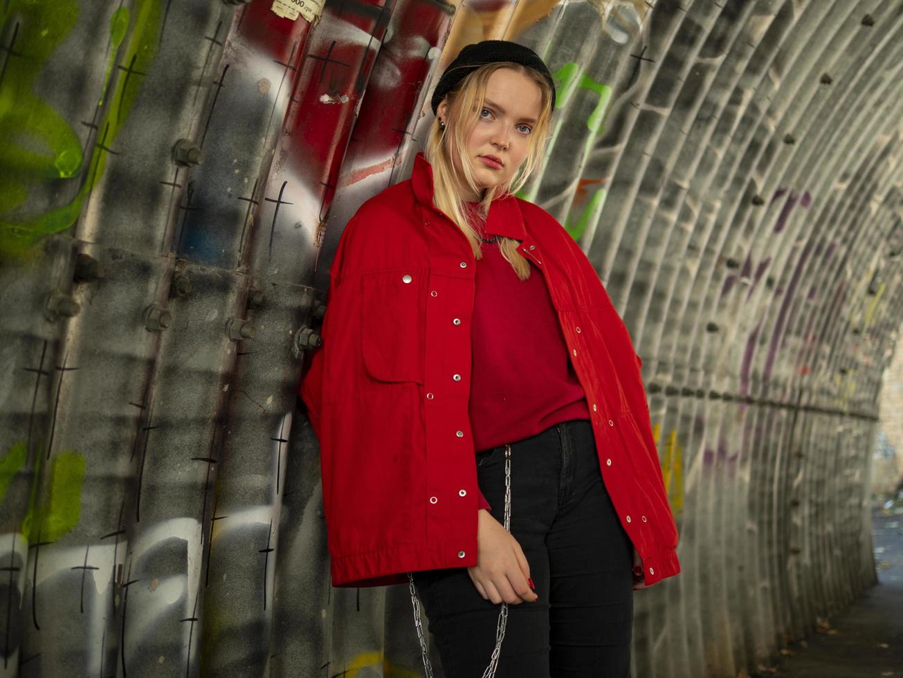 attractive young hipster girl in knitted hat and red jacket is standing under a bridge against the background of a wall with graffiti photo