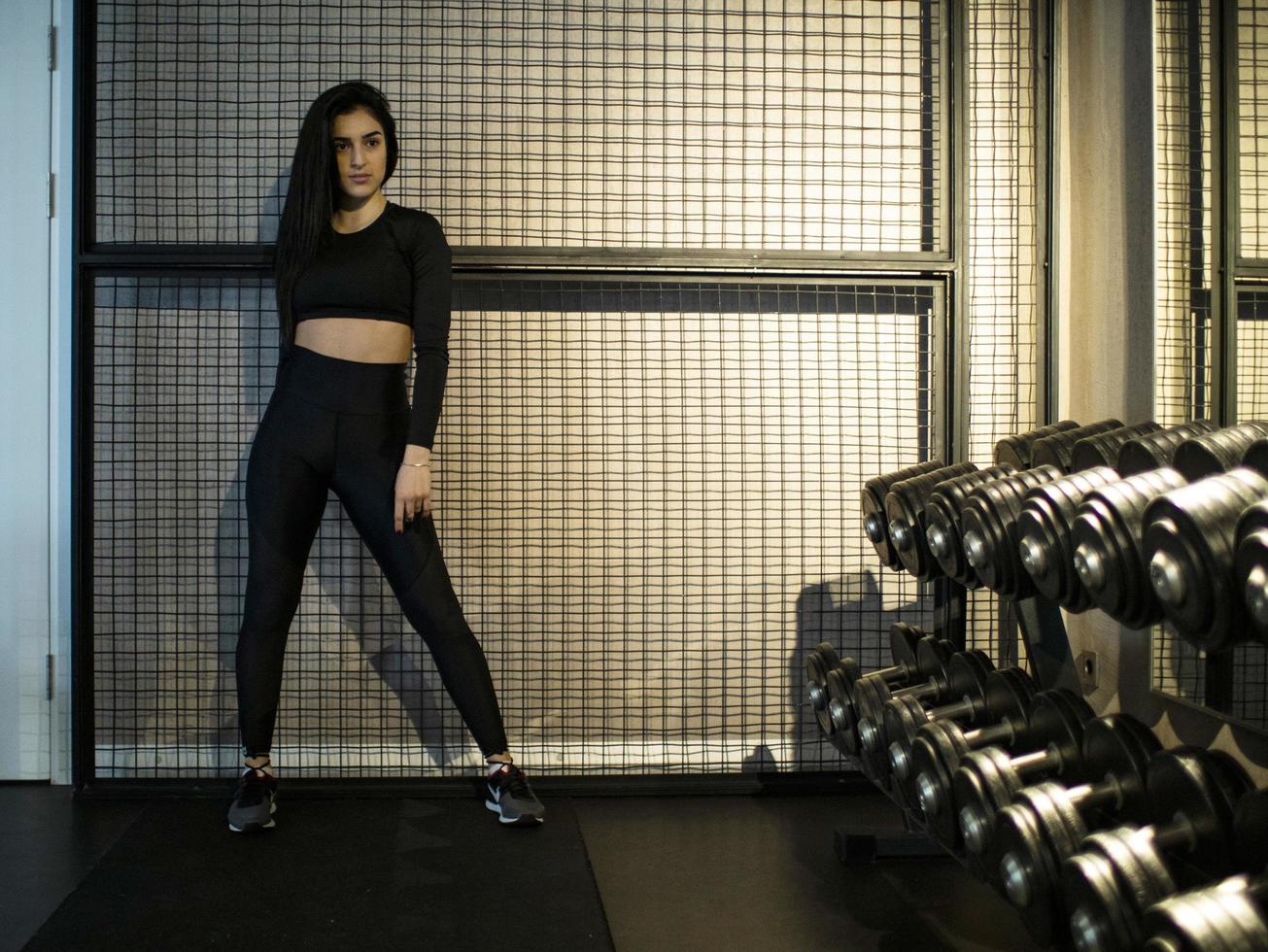 sporty brunette in black leggings and top is standing tall on a shelf with dumbbells in the gym against the background of the lattice photo