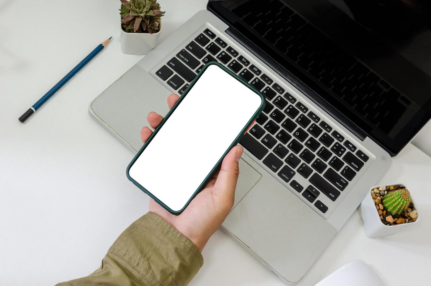 Top view mock up hand holding smart phone white blank screen and pencil mouse computer laptop flat lay on desk photo