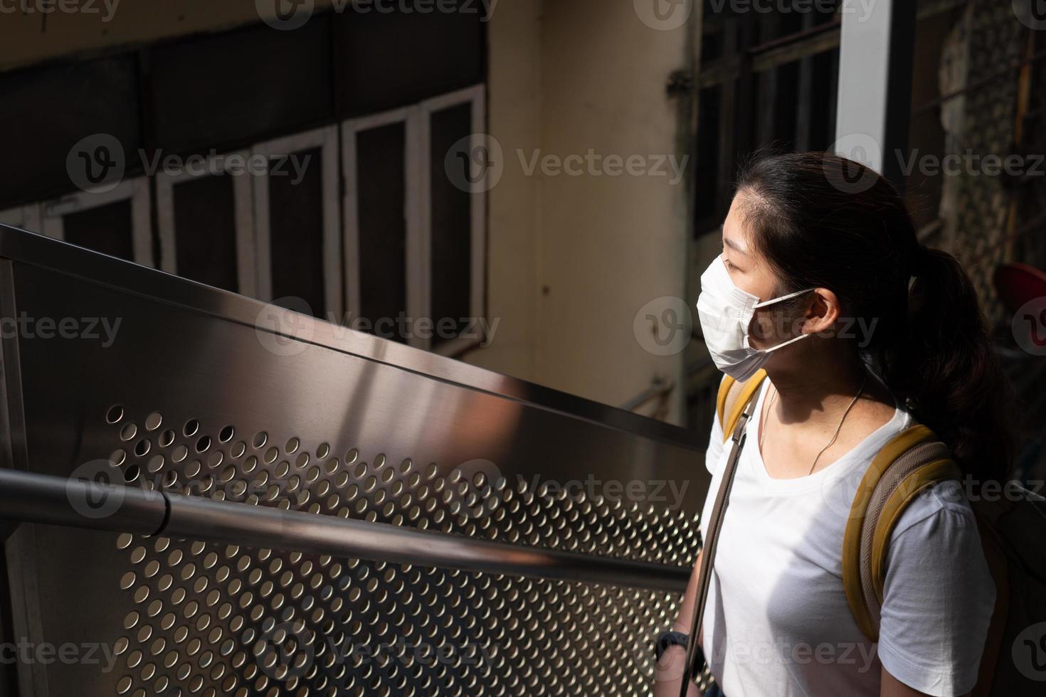 joven y bella mujer asiática con mascarilla como pauta de distanciamiento social. ella está usando la escalera mecánica para subir a la estación de tren del cielo. nuevo estilo de vida normal, covid-19, concepto de coronavirus foto