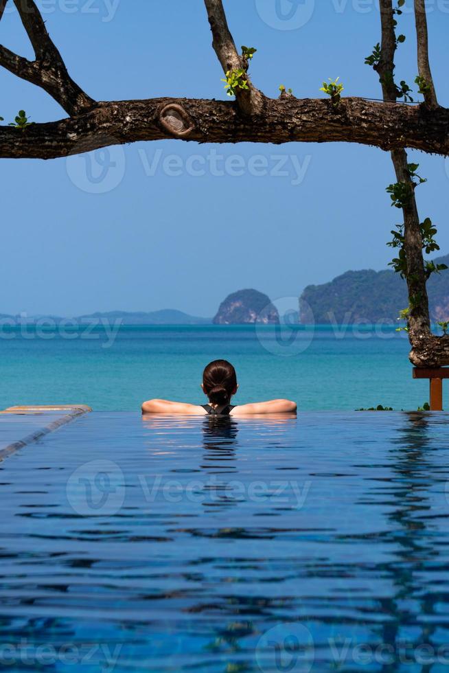 Portrait of young woman wearing bikini standing in the blue infinity swimming pool looking at the view of ocian on summer vacation photo