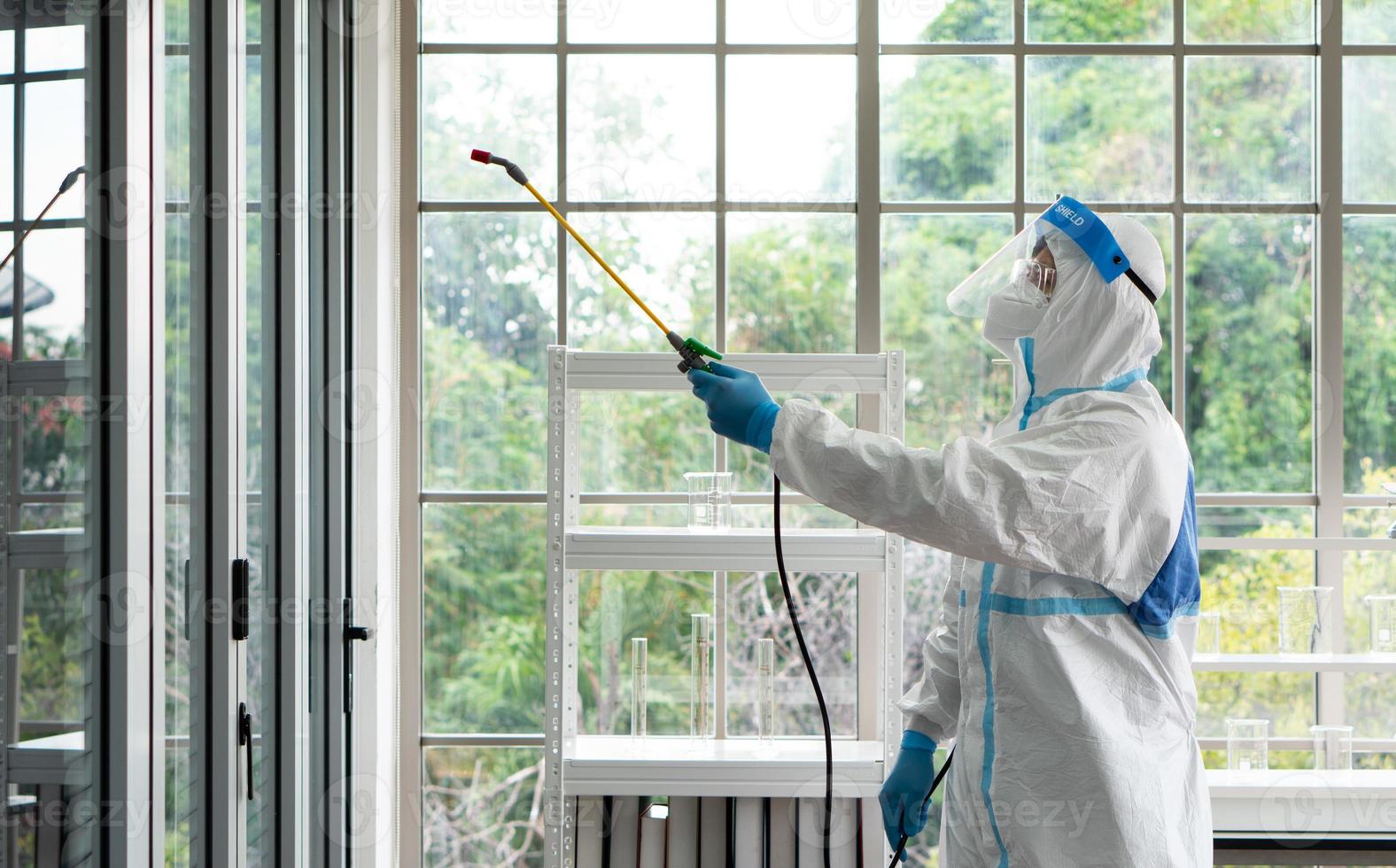 Worker from decontamination services wearing personal protective equipment or ppe including suit, face shield and mask. He uses disinfectant to spray and clean scientist lab photo