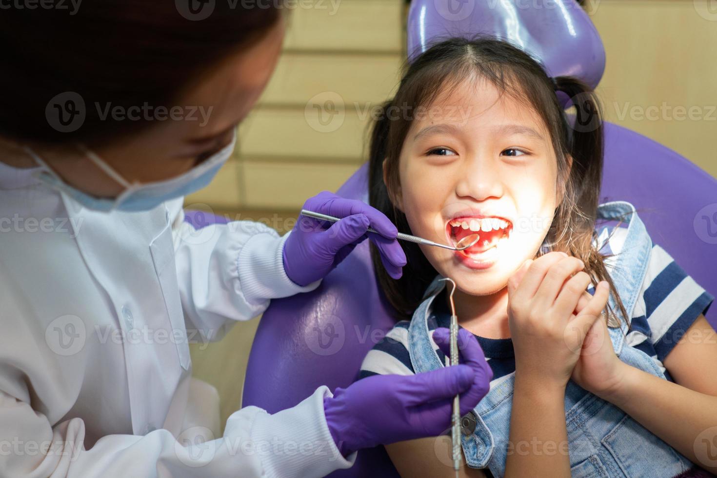 dentista que usa herramientas dentales para limpiar los dientes de un niño asiático y tratar la caries dental en la clínica con ayuda de pie detrás del paciente. concepto de odontología y salud foto