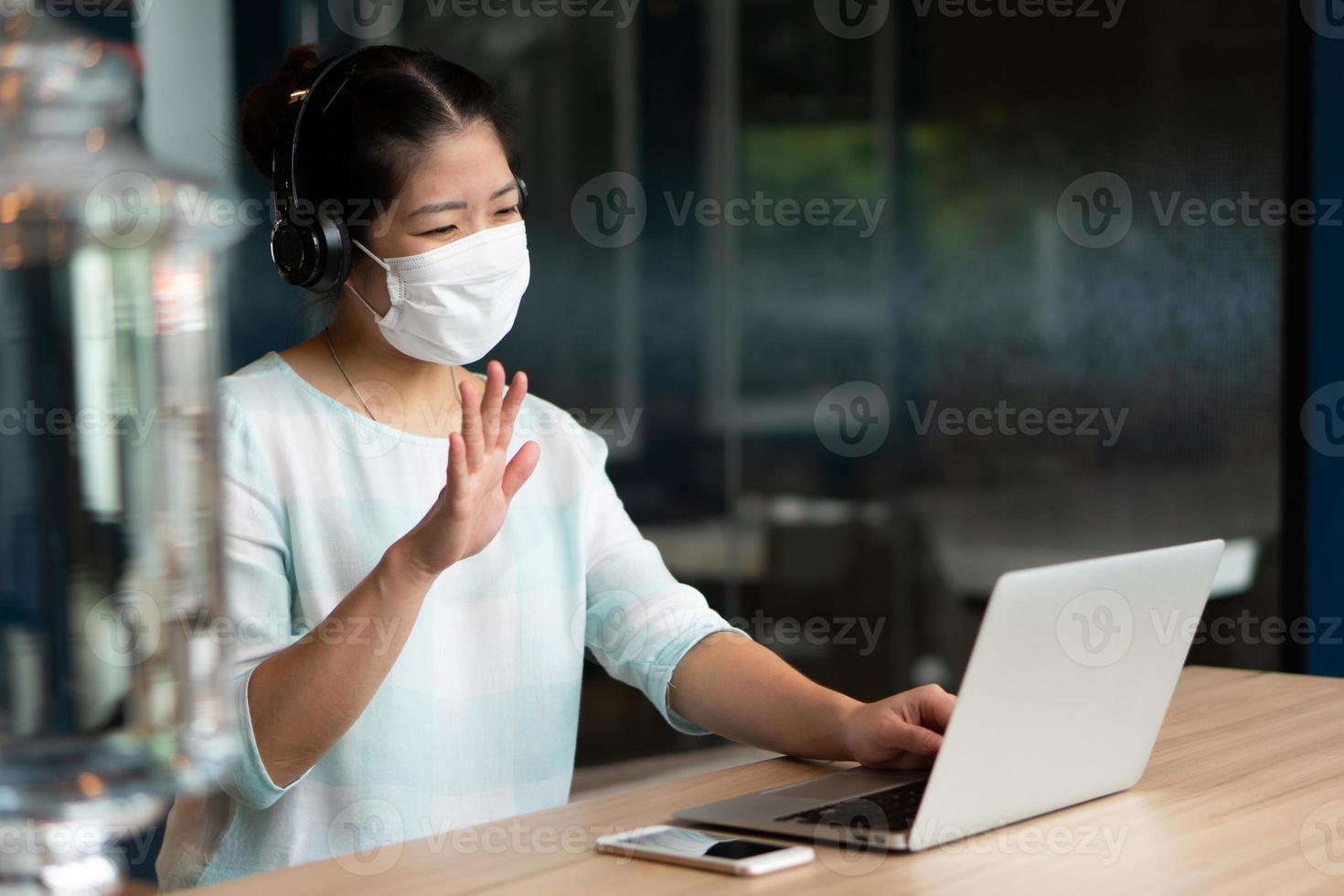 Retrato de mujer asiática joven con mascarilla y auriculares y trabajando de forma remota mediante el uso de conferencias de videollamadas de computadora en el espacio de coworking. distanciamiento social y nuevo concepto de estilo de vida normal foto