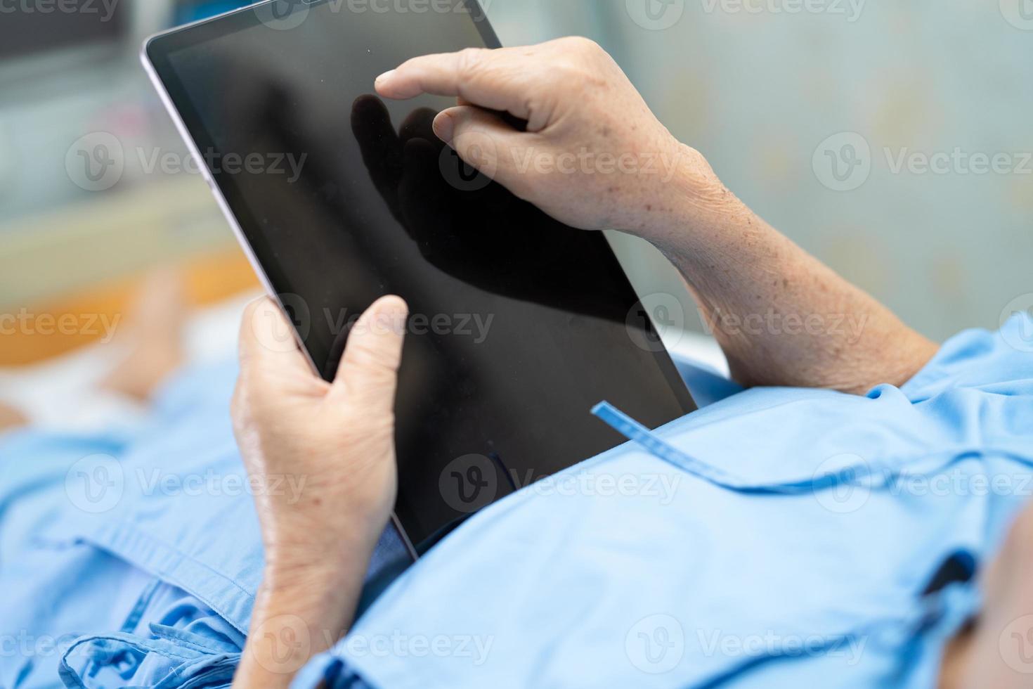 Paciente asiático mayor o anciano mujer sosteniendo en sus manos tableta digital y leyendo correos electrónicos mientras está sentado en la cama en la sala del hospital de enfermería, concepto médico fuerte y saludable foto