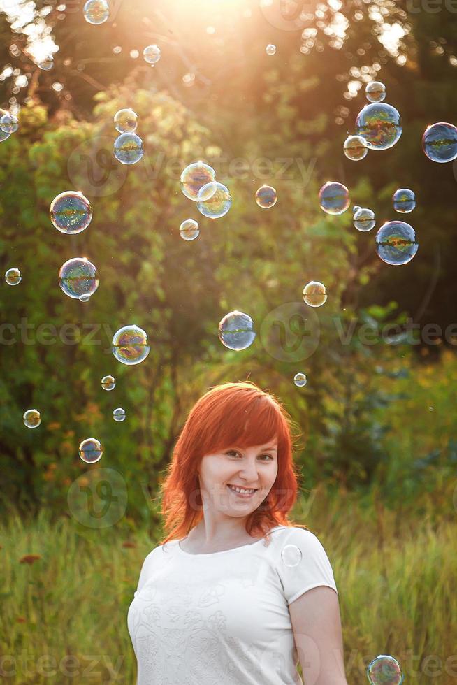 Young woman in a white T-shirt is playing with soap bubbles. Happy red-haired girl in the sun. photo