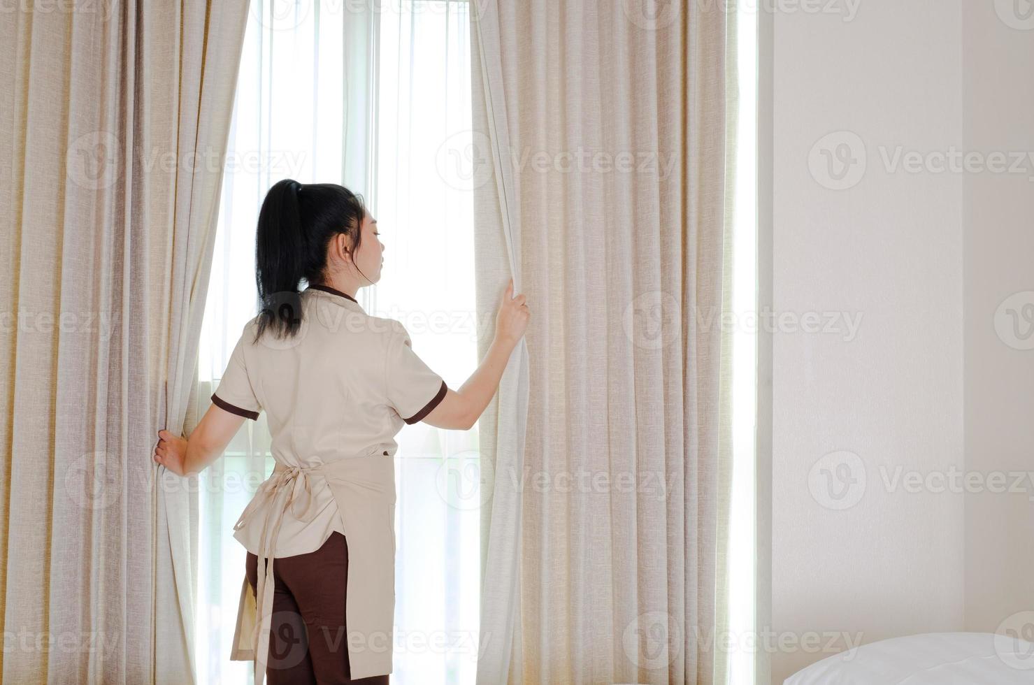 Joven sirvienta abriendo cortinas en la habitación del hotel foto