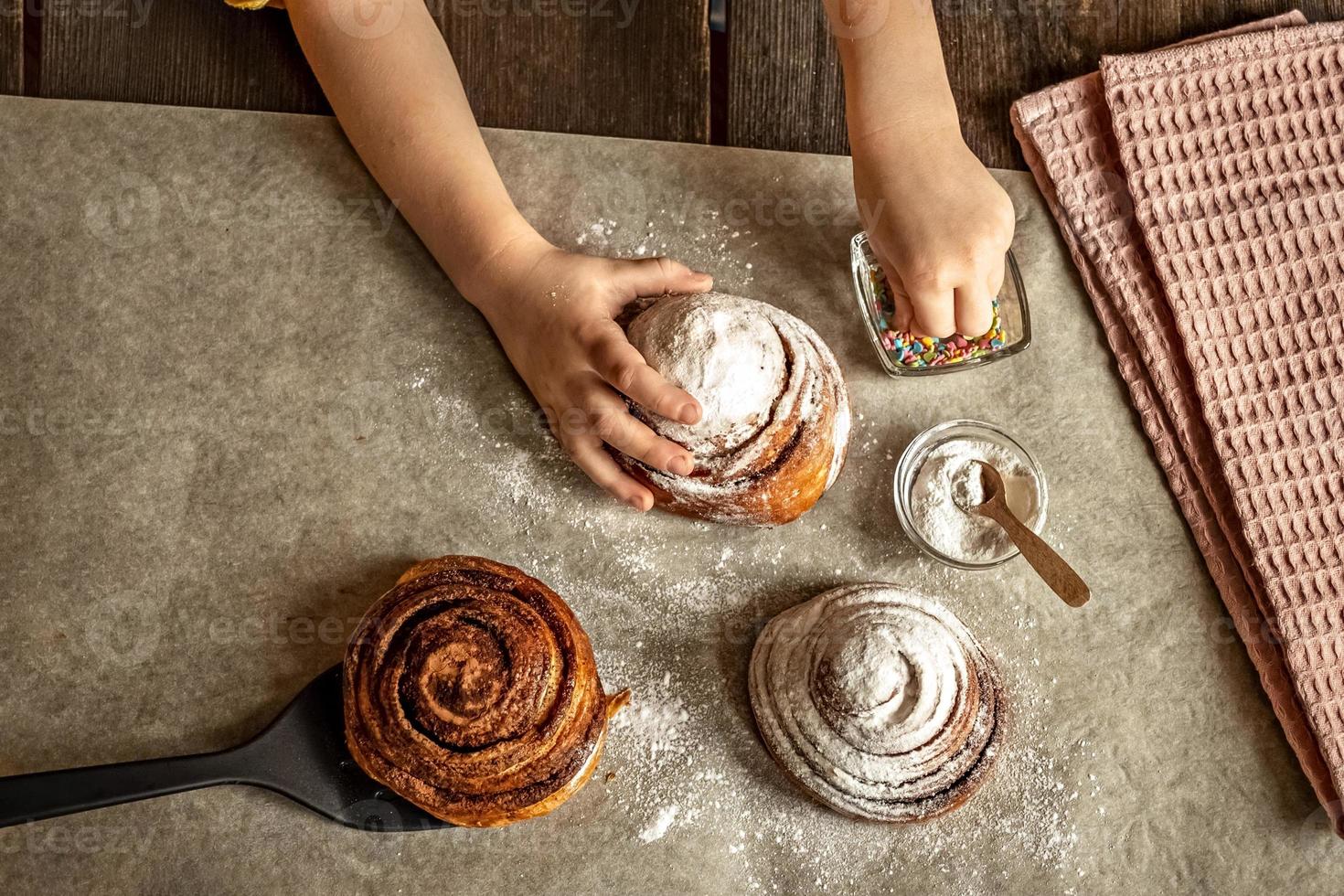 las manos de los niños en el marco espolvorean un bollo recién horneado con canela con chispas de pastelería foto