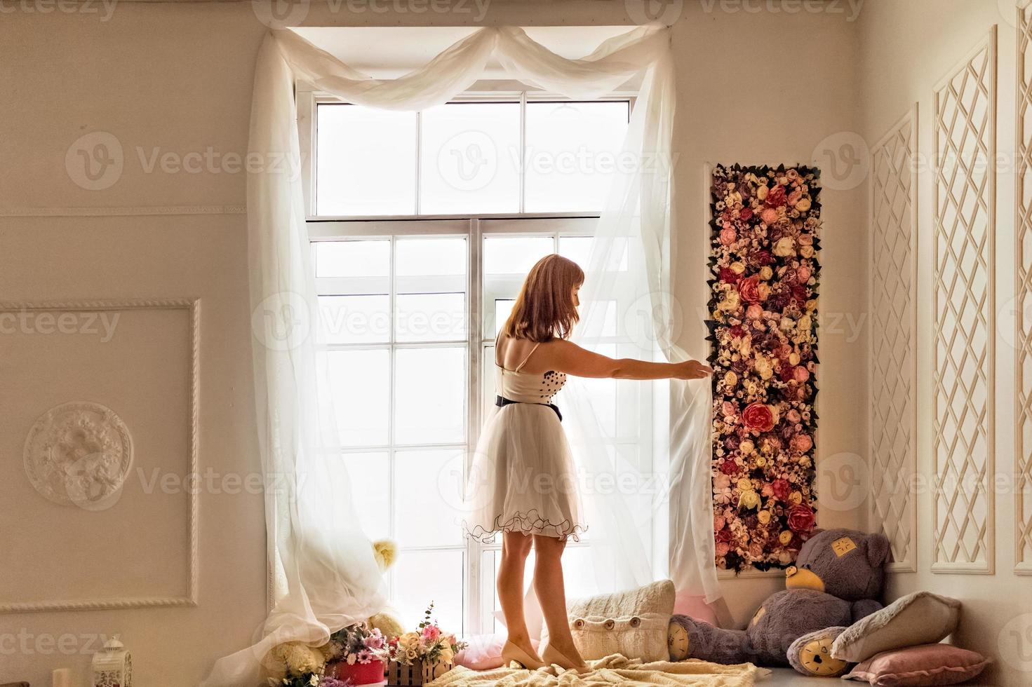 retrato de una mujer joven con un vestido blanco enderezar cortinas blancas claras junto a la ventana. foto