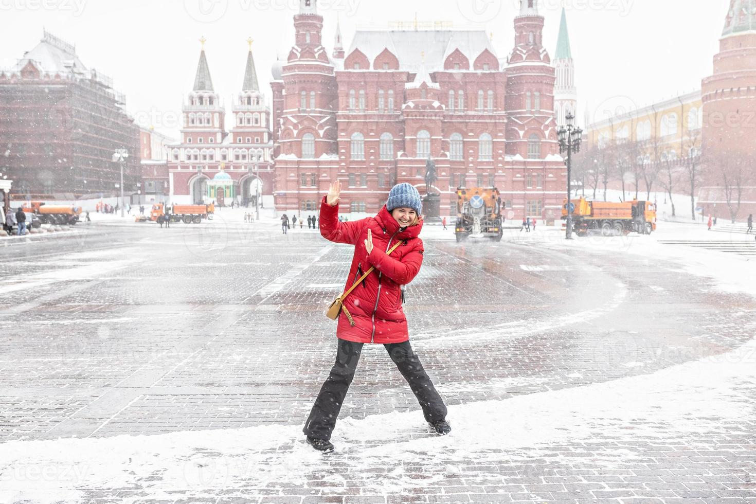 A beautiful young girl in a red jacket walks along Manezhnaya Square in Moscow during a snowfall and blizzard. Snowblowers are working in the background. photo