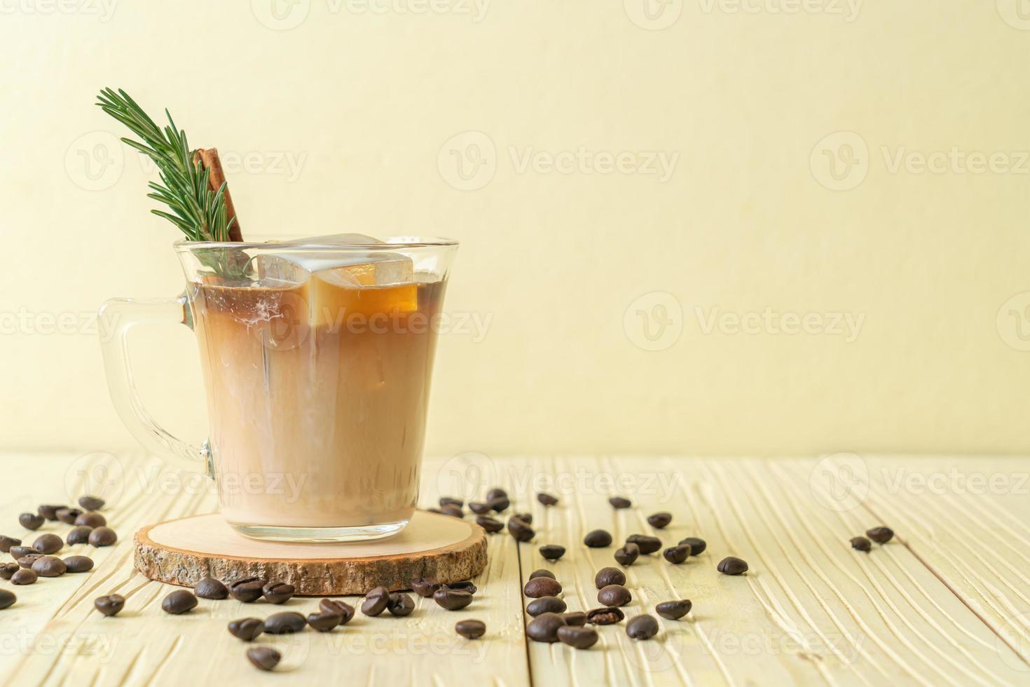 Pouring milk in black coffee glass with ice cube, cinnamon and rosemary on wood background photo