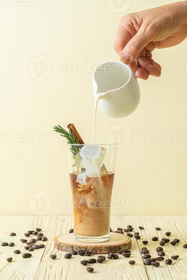 Pouring milk in black coffee glass with ice cube, cinnamon and rosemary on wood background photo