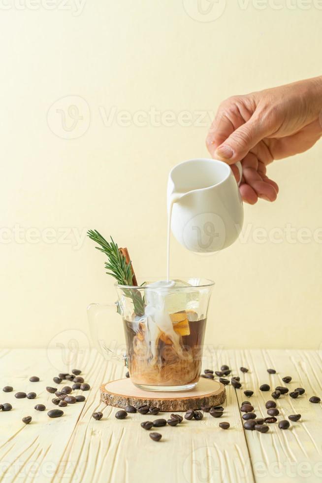 Pouring milk in black coffee glass with ice cube, cinnamon and rosemary on wood background photo