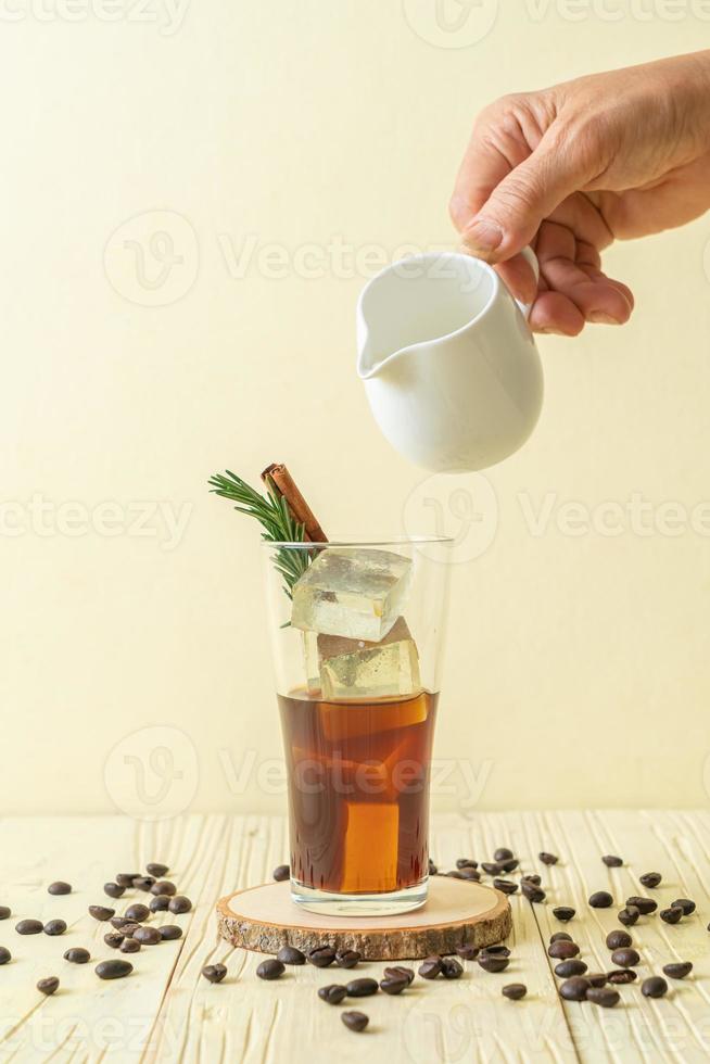 Pouring milk in black coffee glass with ice cube, cinnamon and rosemary on wood background photo