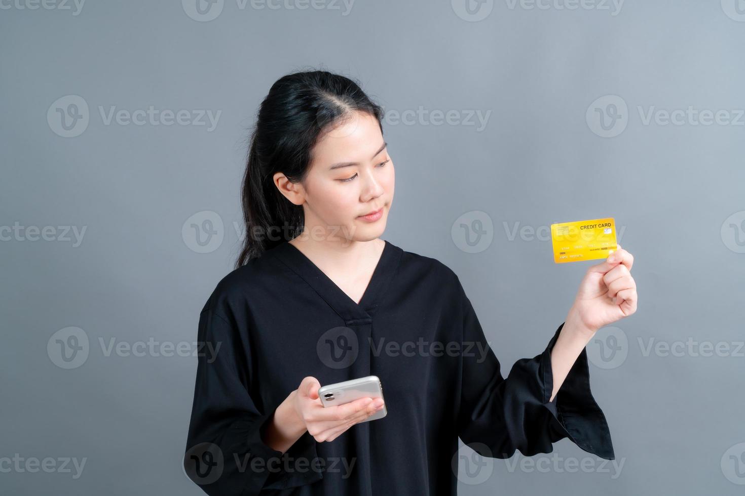 Retrato de una joven asiática feliz mostrando una tarjeta de crédito de plástico mientras sostiene el teléfono móvil sobre fondo gris foto