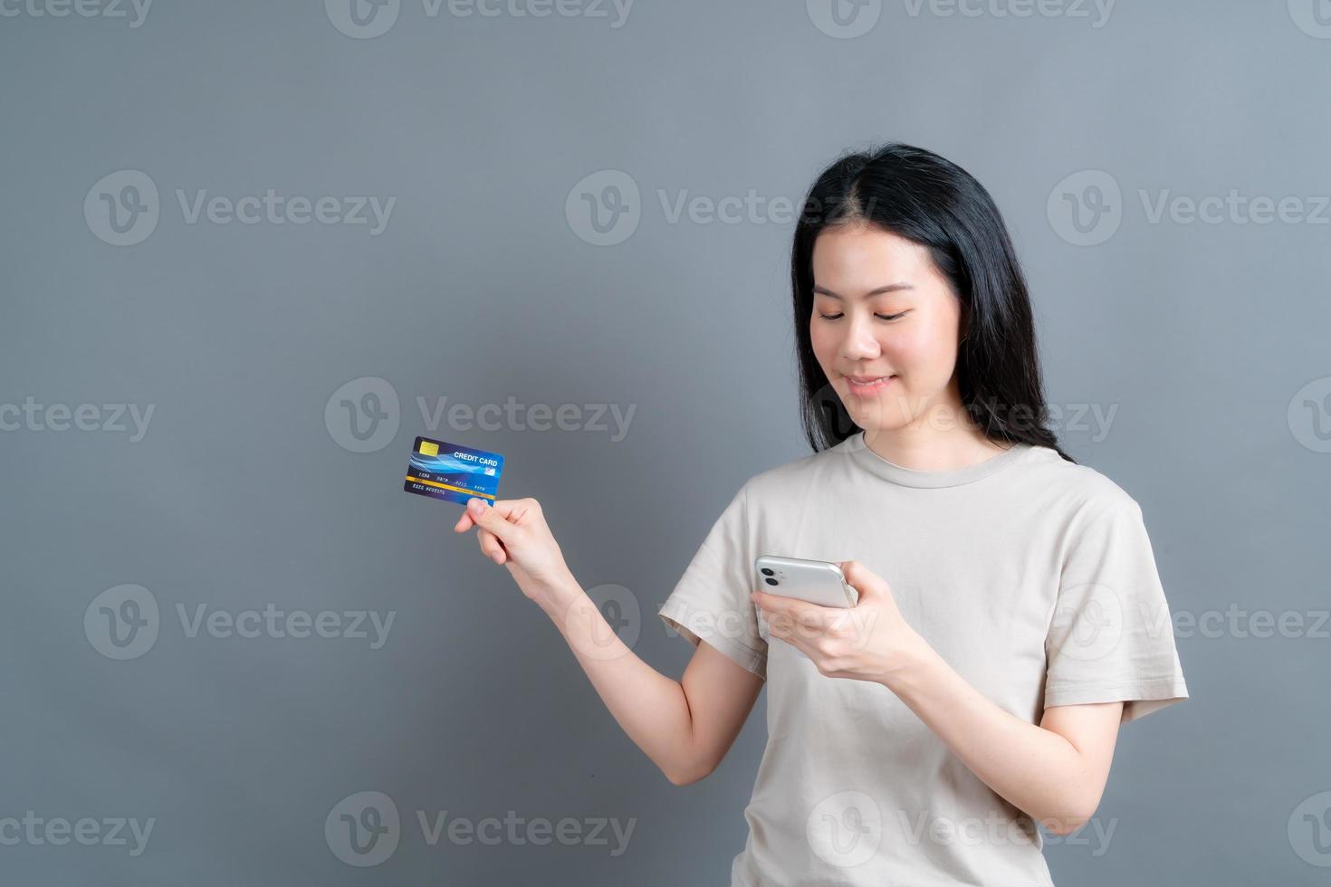 Retrato de una joven asiática feliz mostrando una tarjeta de crédito de plástico mientras sostiene el teléfono móvil sobre fondo gris foto