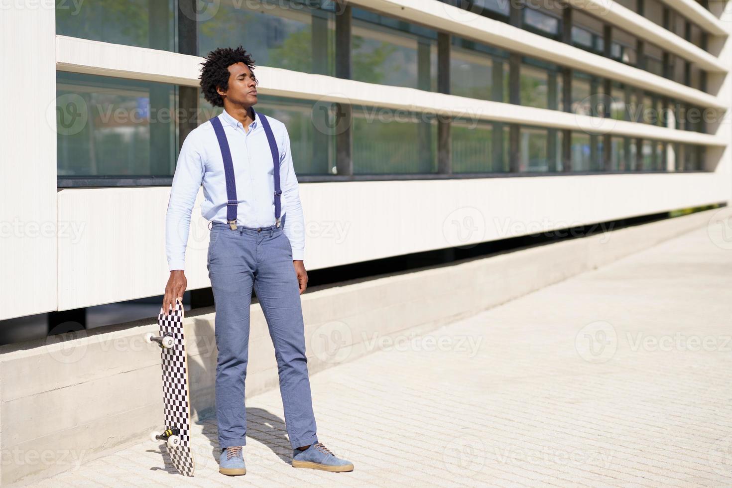 Black male standing next to an office building with a skateboard. photo