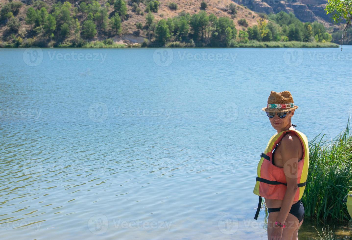 Mujer con sombrero y chaleco salvavidas de pie junto al río. foto