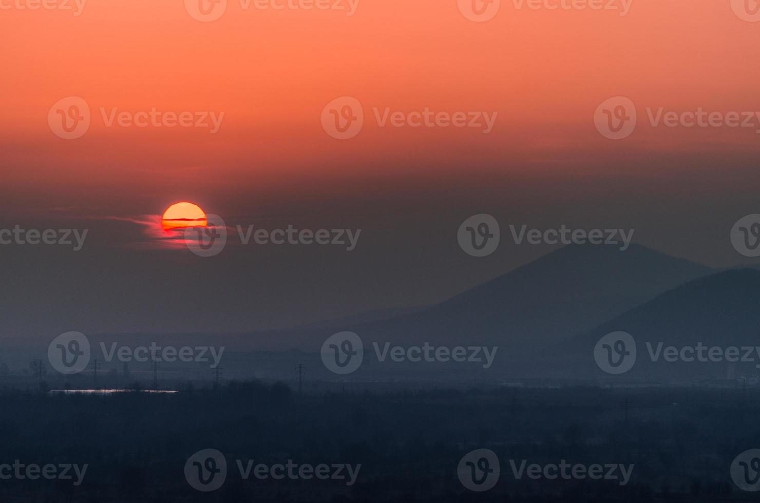 Sunset with silhouettes of hills in background, sun covered by clouds photo