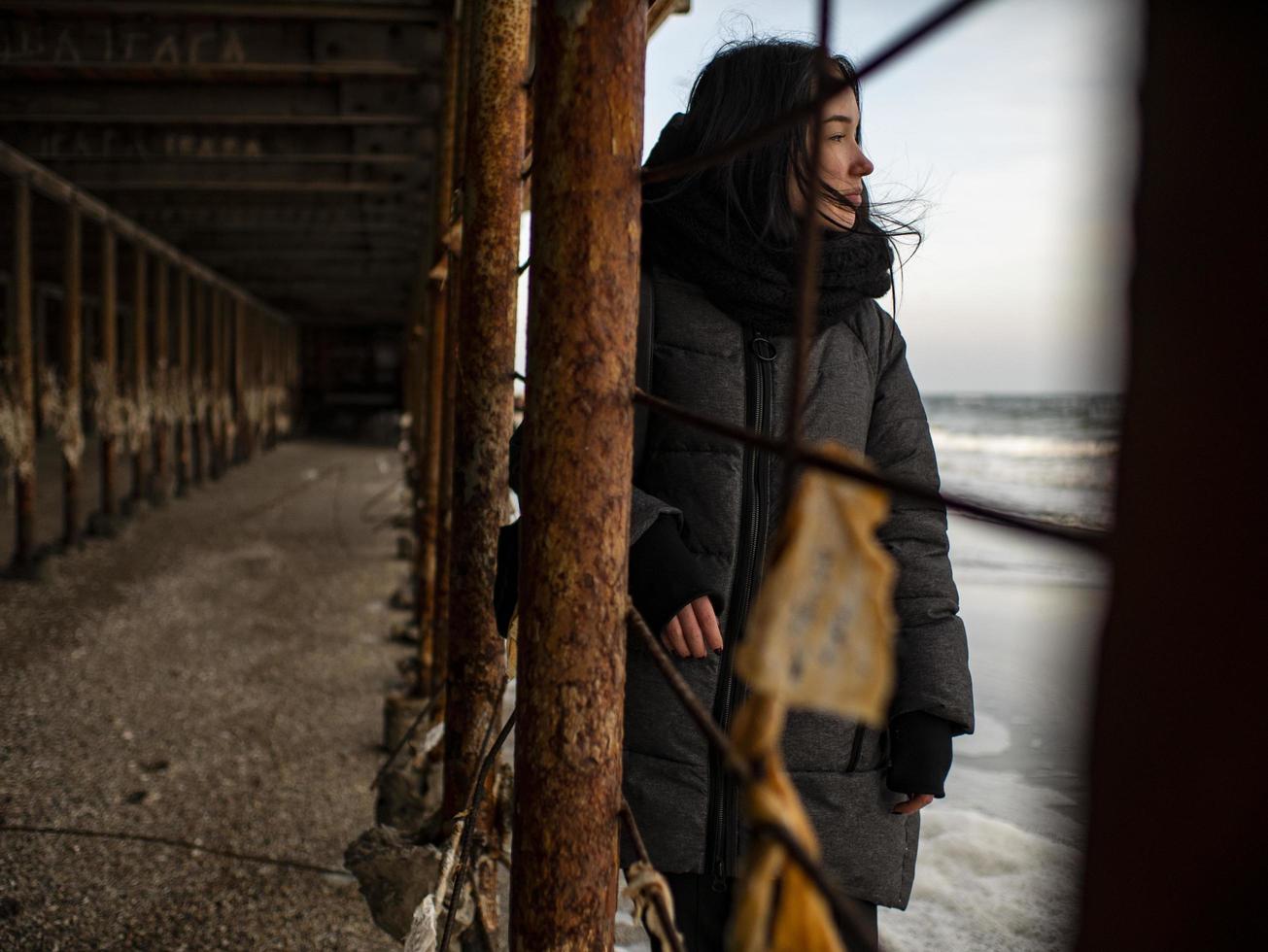 young girl in a coat stands near metal rusty structures near the sea photo