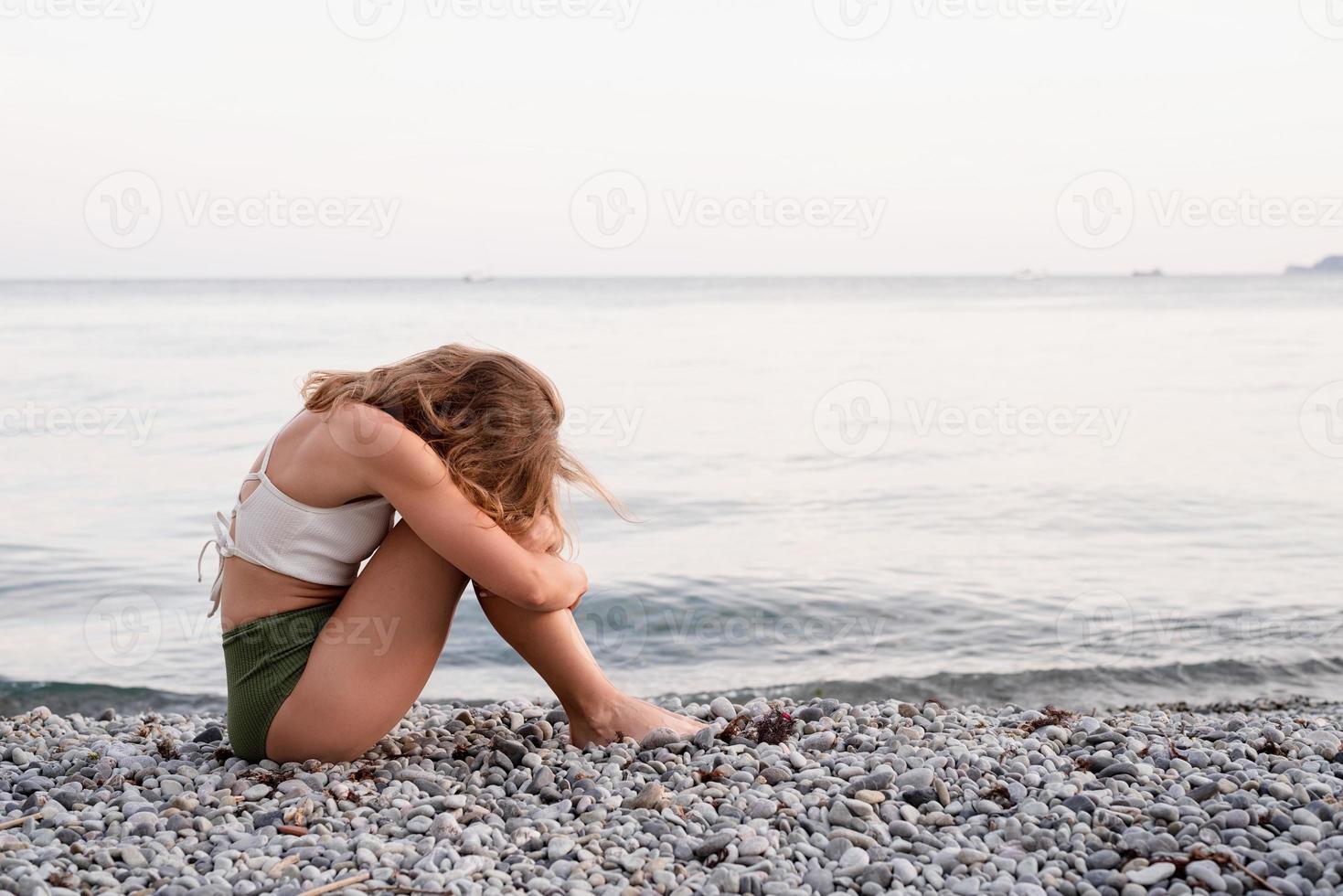 Joven mujer deprimida sentada en la playa mirando a otro lado, vista trasera foto