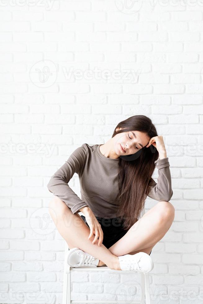 Mujer con cabello largo sentado sobre fondo de pared de ladrillo blanco, pensando foto