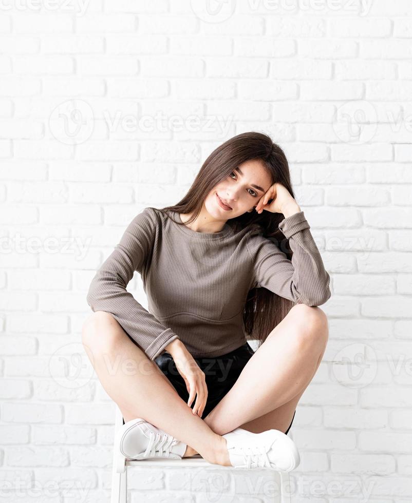 Mujer con cabello largo sentado sobre fondo de pared de ladrillo blanco foto