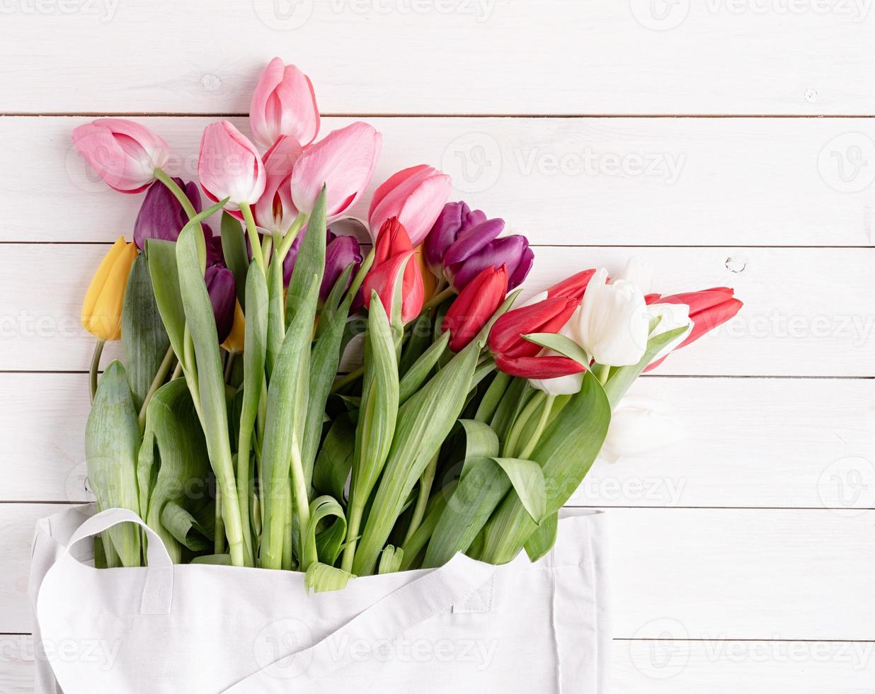 Bolsa de tela gris llena de coloridos tulipanes sobre fondo blanco de madera foto