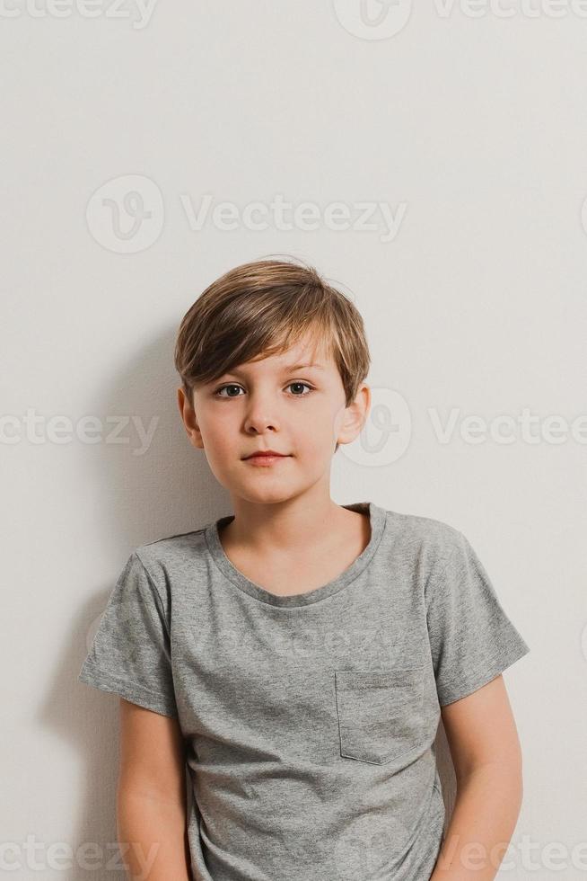 A cute boy standing next to white wall, grey shirt, slightly smiling photo