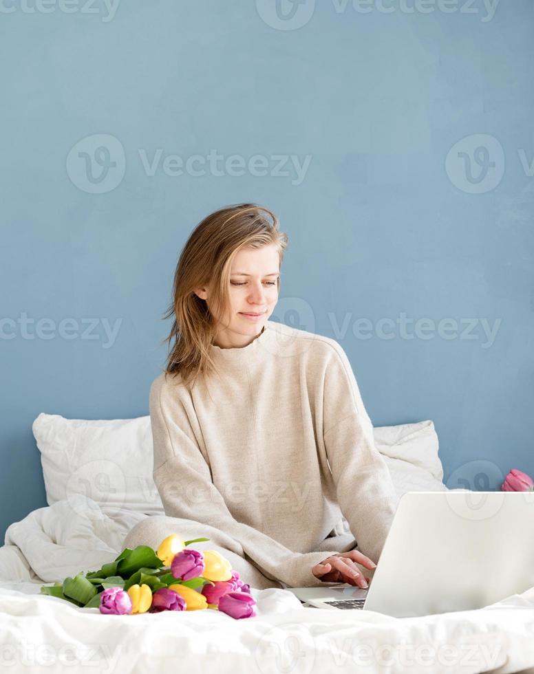 Feliz mujer sentada en la cama en pijama trabajando en un portátil foto