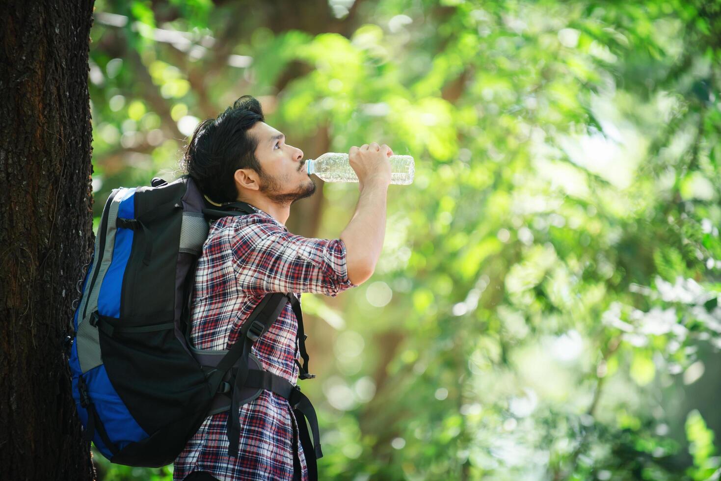 joven sediento y beber agua durante la caminata detrás de un gran árbol. foto