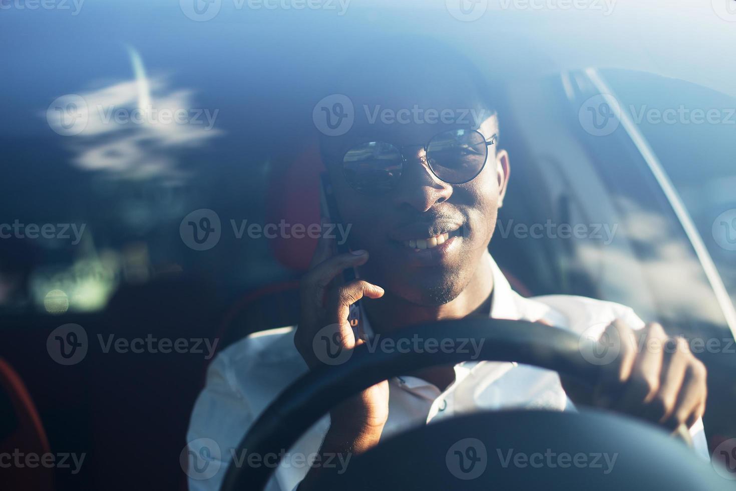 Feliz afroamericano conduciendo un coche con un teléfono, en el verano foto
