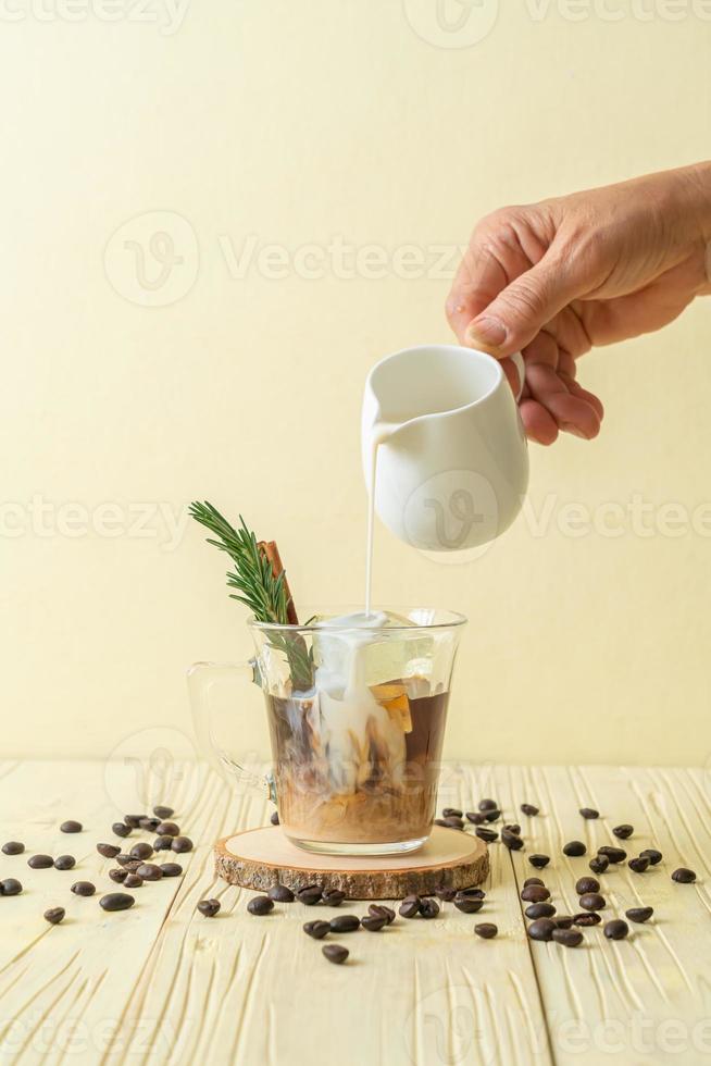 Pouring milk in black coffee glass with ice cube, cinnamon, and rosemary on wood background photo